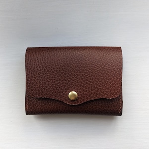 May include: A small, brown leather wallet with a textured surface. The wallet has a flap closure secured by a gold-colored button. The wallet is rectangular and appears to be new.