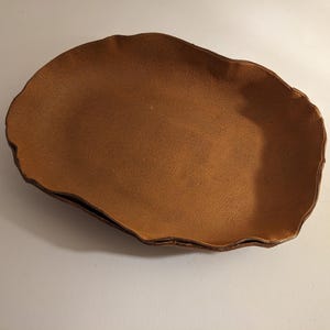 May include: A brown, irregularly shaped leather bowl. The bowl has a textured surface and a dark brown edge. The bowl is empty and sits on a white surface. The bowl is likely handmade.