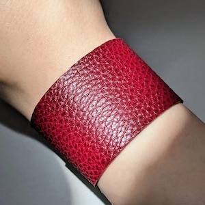 May include: A wide, red leather cuff bracelet with a textured surface. The bracelet is a deep shade of red and appears to be made of genuine leather. The cuff is approximately 5 cm wide.