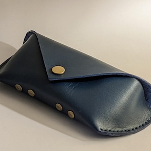 May include: A navy blue leather glasses case with a triangular flap secured by a brass button. The case has a rounded bottom and is secured with three additional brass rivets. The stitching is visible along the edges.