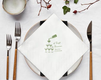 May include: A white square napkin featuring a green illustration of champagne being poured into a pyramid of glasses. The text reads "A Toast to Forever Selena + Connor". The napkin is on a plate with cutlery.