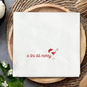 May include: White square paper napkin with a diamond pattern. The napkin features red text that reads "a tini bit merry" and a graphic of a cocktail with a Santa hat and candy cane.