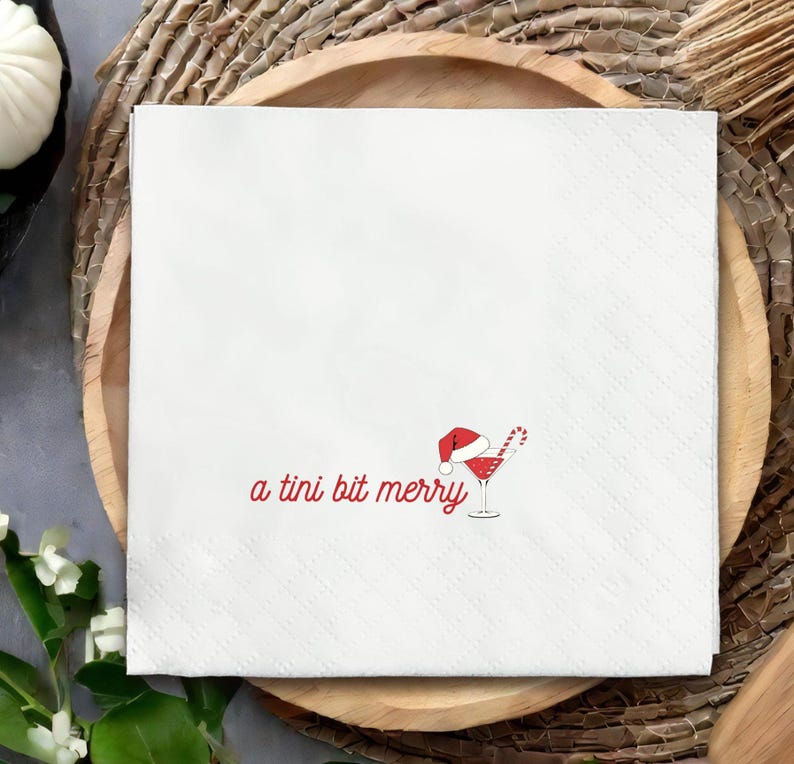 May include: White square paper napkin with a diamond pattern. The napkin features red text that reads "a tini bit merry" and a graphic of a cocktail with a Santa hat and candy cane.