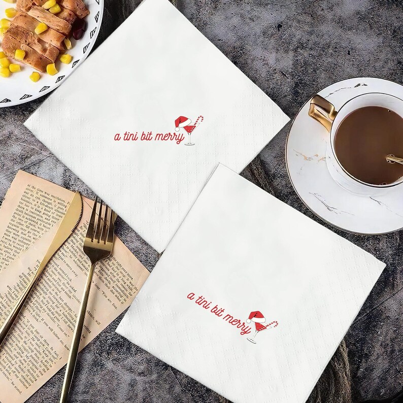 May include: Two white cloth napkins with the phrase "a tini bit merry" embroidered in red, along with a Santa hat and cocktail glass design. The napkins are on a dark gray surface, near a cup of coffee and a plate of food.