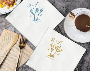 May include: Two white cocktail napkins with custom printing. One napkin has blue text and a cocktail glass design with the initials "R+L" and the date "15.12.2025". The other napkin has gold text and a similar design.