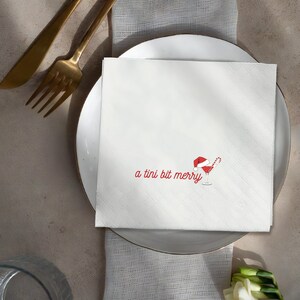 May include: White square napkin with a diagonal textured pattern. The napkin features the phrase "a tini bit merry" in red script, along with a red cocktail glass with a Santa hat and candy cane.