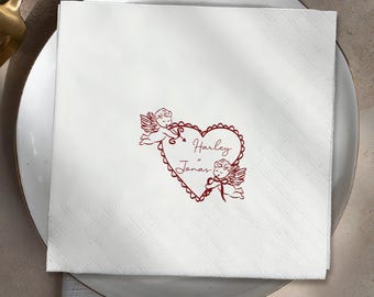 May include: White square paper napkin with a red heart design, featuring two cherubs and the names "Harley Jonas". The napkin is placed on a white plate, with a gold-colored utensil visible in the background. The napkin has a subtle diamond pattern.