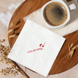 May include: White cocktail napkin with the text "a tini bit merry" in red script. A red Santa hat and candy cane are next to a martini glass illustration. A cup of coffee sits on a wooden tray.