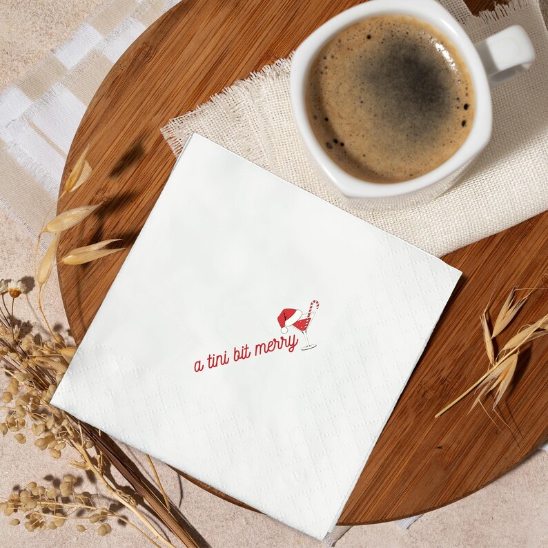 May include: White cocktail napkin with the text "a tini bit merry" in red script. A red Santa hat and candy cane are next to a martini glass illustration. A cup of coffee sits on a wooden tray.