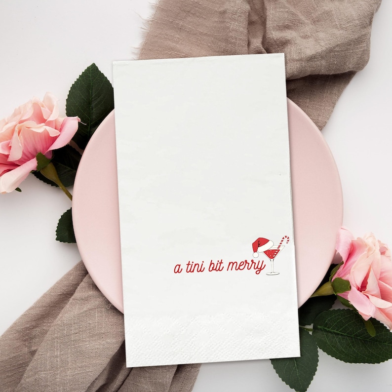 May include: White rectangular napkin with the phrase "a tini bit merry" in red script, along with a Santa hat, candy cane, and cocktail illustration. The napkin is on a pink plate, with pink flowers and a tan cloth in the background.