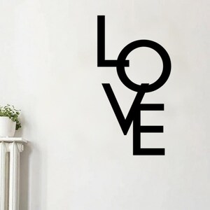 Könnte beinhalten: Schwarze Metall-Wandkunst mit dem Wort "LOVE" in einer modernen, minimalistischen Schriftart. Das Kunstwerk ist an einer weißen Wand über einem hellblauen Sofa angebracht. Der Raum ist mit Pflanzen und einem Beistelltisch aus Holz dekoriert.