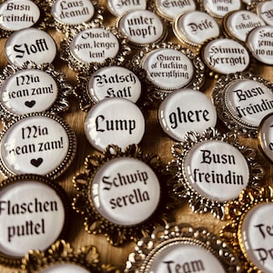 Könnte beinhalten: Eine Sammlung verzierter Anstecknadeln im Vintage-Stil mit weißen Zentren und gold- oder silberfarbenen Filigranrahmen. Jede Nadel zeigt einen anderen deutschen Satz in schwarzer Schrift, wie "Queen of everything" und "Busn freindin".
