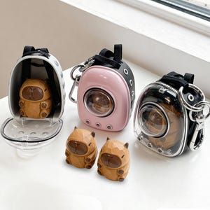 May include: Three miniature pet carrier keychains in white, pink, and black, each with a clear viewing window. Two small, brown capybara figurines sit in front of the carriers. The carriers have black straps and silver-colored hardware.
