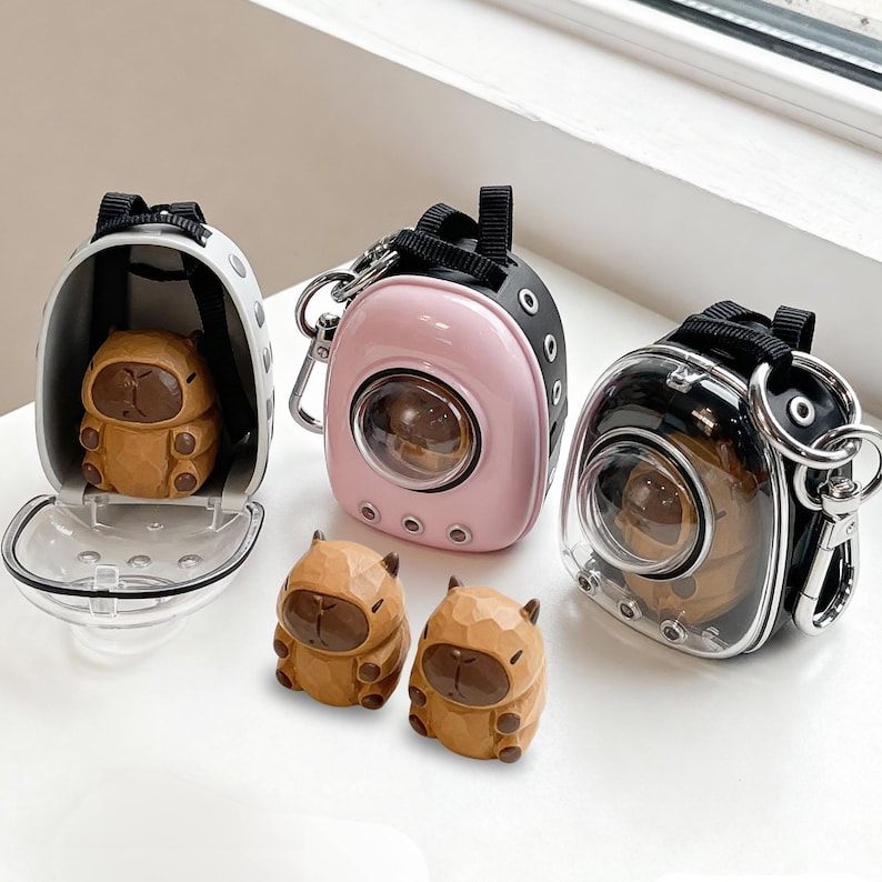 May include: Three miniature pet carrier keychains in white, pink, and black, each with a clear viewing window. Two small, brown capybara figurines sit in front of the carriers. The carriers have black straps and silver-colored hardware.