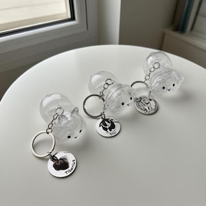 May include: Clear plastic keychains with cartoon animal faces and silver keyrings. Each keychain has a small, round charm with a black and white animal illustration and a name. The keychains are on a white surface.