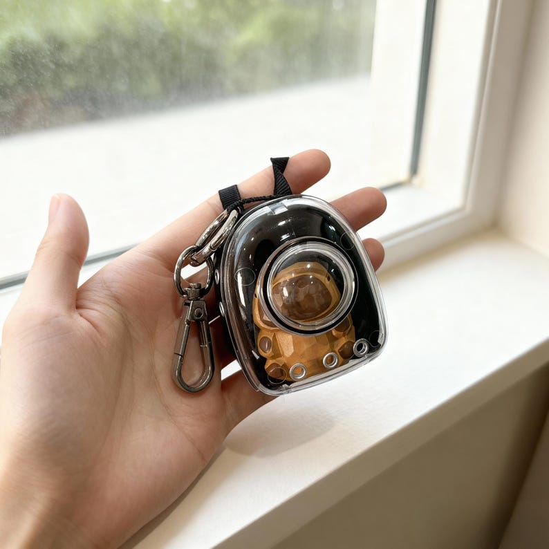 May include: A clear, black, and silver keychain featuring a small astronaut-themed carrier. The carrier has a clear bubble window, and a small, brown astronaut figure is visible inside. The keychain includes a black strap and a silver clasp.