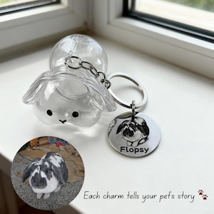 May include: A clear plastic bunny-shaped keychain with a silver keyring and a circular tag. The tag features a black and white illustration of a rabbit and the name "Flopsy". The text "Each charm tells your pet's story" is also visible.