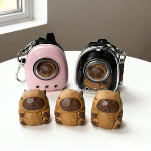 May include: Two small pet carriers, one pink and one black, with clear viewing windows and silver hardware. Three carved wooden capybara figurines are in front of the carriers. The carriers have a handle and a clip for attaching to a bag.