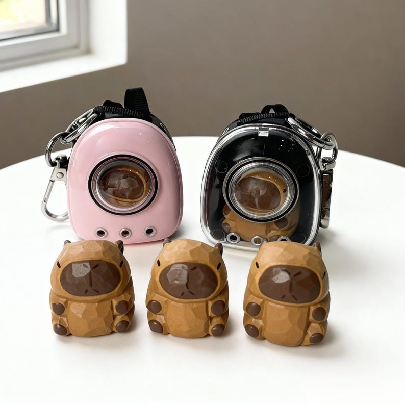 May include: Two small pet carriers, one pink and one black, with clear viewing windows and silver hardware. Three carved wooden capybara figurines are in front of the carriers. The carriers have a handle and a clip for attaching to a bag.