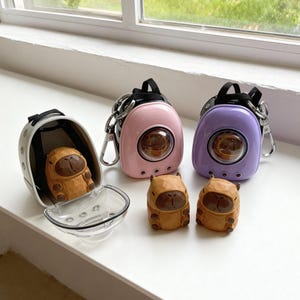 May include: A collection of capybara-themed accessories. Includes a gray carrier with a clear front, two backpack-style keychains in pink and purple, and three wooden capybara figurines. The keychains have a circular window.
