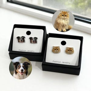 May include: Two pairs of stud earrings in black boxes. One pair features dog faces, the other cat faces. The dog earrings are black, white, and brown. The cat earrings are orange and white. Each pair is displayed on a white card with "Fashion Jewelry" printed on it.