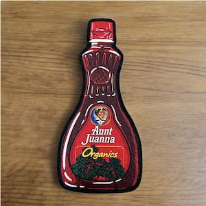 May include: Aunt Juanna Organics syrup bottle-shaped patch. The patch is dark red with a white border and features the Aunt Juanna logo and the word "Organics". The patch is on a wooden surface.