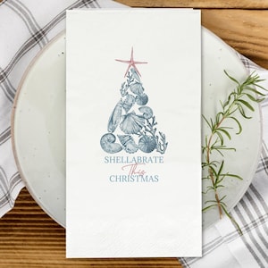 May include: A white paper napkin featuring a printed graphic of a Christmas tree constructed from various blue seashells and coral, topped with a pink starfish. The text below the tree reads, "SHELLABRATE This CHRISTMAS" in blue and pink lettering.