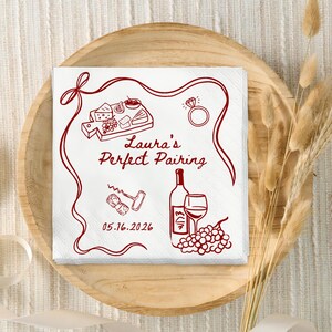 May include: White square paper napkins with a red illustration of wine, cheese, and a ring. The text "Laura's Perfect Pairing" and the date 05.16.2026 are also printed on the napkins. The napkins are on a wooden plate.