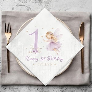 May include: A white paper napkin featuring a large purple number one, a fairy illustration with a purple dress and wings, and the text Happy 1st Birthday EVELYN in purple and gold lettering, accented with small gold stars and floral details.