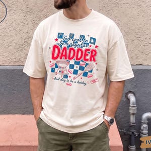 Puede incluir: Camiseta color marfil con el texto "Too cool to be a regular DADDER" en rojo y azul. El diseño incluye una parrilla, una espátula y perritos calientes. La camiseta es de corte holgado.