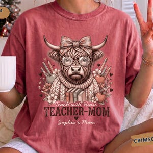 Puede incluir: Una camiseta carmesí con un gráfico de una vaca de las Tierras Altas con gafas y un lazo, rodeada de lápices, manzanas y flores. El texto dice Teach with Heart, TEACHER-MOM y Sophia's Mom. La camiseta tiene cuello redondo y mangas cortas.