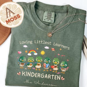 May include: Green Comfort Colors t-shirt with the text "Loving Littlest Learners KINDERGARTEN Mrs. Anderson" and a graphic of cartoon ducks. A gold necklace with a pendant is draped across the shirt.
