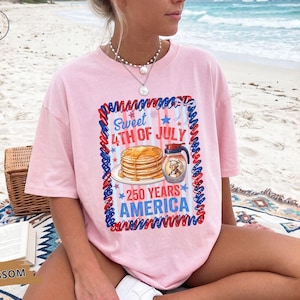 Puede incluir: Una camiseta rosa claro con un estampado gráfico de una pila de tortitas y una jarra de sirope. El texto dice Sweet 4th of July 250 Years America. El diseño está enmarcado por un borde de cuerda trenzada roja, blanca y azul con estrellas.