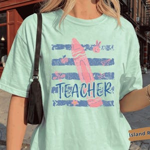 Puede incluir: Una camiseta de color verde menta claro con un estampado gráfico de un crayón rosa centrado sobre rayas horizontales azul marino desgastadas. La palabra TEACHER está impresa en letras mayúsculas azul marino debajo del crayón, rodeada de pequeños iconos rosas.