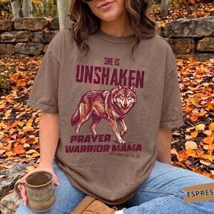 Puede incluir: Una camiseta marrón de manga corta con un gráfico de un lobo en el centro. El texto sobre el lobo dice "SHE IS UNSHAKEN" y el texto debajo dice "PRAYER WARRIOR MAMA" seguido de "PROVERBS 31: 25-26" en color rojo oscuro.