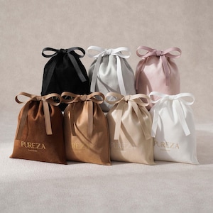 May include: A collection of seven drawstring pouches in black, light gray, dusty pink, dark brown, tan, beige, and white. Each pouch features a matching ribbon tie at the top and the gold text PUREZA PACKAGE printed on the front lower section.
