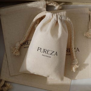 May include: Beige drawstring pouches with the text "PUREZA PACKAGE" printed in black. The pouches have a natural, textured appearance and are made of a soft fabric. The drawstring is a thick, twisted rope.