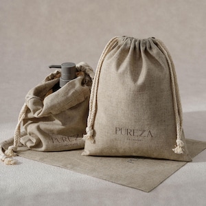 May include: Two beige linen drawstring bags with the text "PUREZA PACKAGE" printed on them. One bag contains a grey pump bottle. The bags are on a matching beige linen mat. The drawstring cords are off-white.