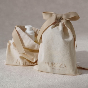 K&ouml;nnte beinhalten: Zwei cremefarbene Baumwollbeutel mit Kordelzug und beigen Schleifen. Auf der Vorderseite der Beutel steht der Text PUREZA PACKAGE in einer Serifenschrift. Eine wei&szlig;e Kunststoff-Pumpflasche ist teilweise im ge&ouml;ffneten Beutel links zu sehen.