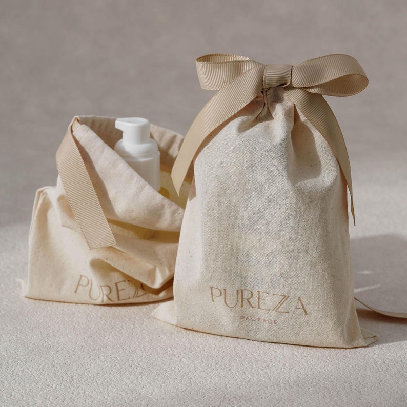 K&ouml;nnte beinhalten: Zwei cremefarbene Baumwollbeutel mit Kordelzug und beigen Schleifen. Auf der Vorderseite der Beutel steht der Text PUREZA PACKAGE in einer Serifenschrift. Eine wei&szlig;e Kunststoff-Pumpflasche ist teilweise im ge&ouml;ffneten Beutel links zu sehen.