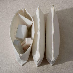 May include: An open white canvas tote bag filled with white boxes and containers, next to two closed white pillow-shaped bags. The bags are on a textured, off-white surface. The tote bag has a simple design.