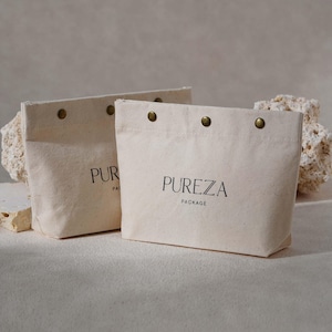 May include: Two cream-coloured canvas tote bags with brass rivets along the top edge. The bags have the word "PUREZA" printed in black, with "PACKAGE" below. The bags are on a light-coloured surface.