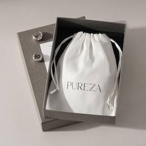 Pode incluir: Uma caixa de presente retangular cinza contendo uma bolsa branca com cordão e o texto PUREZA impresso em preto. Dois pequenos brincos de argola prateados estão colocados no lado esquerdo da caixa, sobre um inserto de cartão branco.