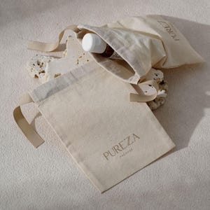 May include: Two beige canvas drawstring bags with the text "PUREZA PACKAGE" printed in a light brown color. One bag is open, revealing a small brown bottle with a white cap. The bags are on a textured, off-white surface.