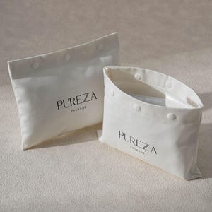 May include: Two off-white fabric pouches with snap closures. The larger pouch is closed, while the smaller one is open, revealing a clear interior. Both pouches have the word "PUREZA" printed in black, with "PACKAGE" below.