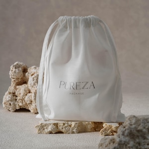 Peut inclure: Un sac blanc à cordon de serrage avec le texte "PUREZA PACKAGE" imprimé en gris. Le sac est posé sur une surface de pierre claire et texturée, avec d'autres pierres en arrière-plan. Le sac est fait d'un matériau doux et translucide.