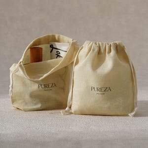 May include: Two off-white drawstring pouches made of fabric, each featuring the black text PUREZA PACKAGE printed on the front. One pouch is closed, while the other is open, revealing a wooden cylinder and a white card with black text inside.