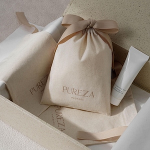 Puede incluir: Un paquete de cuidado de la piel con una bolsa beige con cordón con el texto dorado "PUREZA PACKAGE". También se ve un tubo blanco de espuma limpiadora y una botella. Los artículos están colocados en una caja beige.