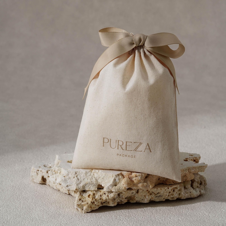 May include: A beige drawstring bag with the word "PUREZA" printed on the front, tied with a matching ribbon. The bag is sitting on a textured, light-colored stone surface. The word "PACKAGE" is printed below "PUREZA".
