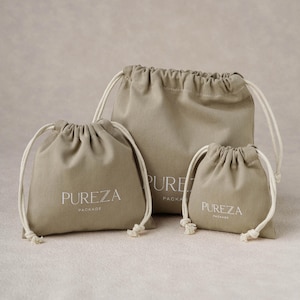 May include: Three beige drawstring pouches of varying sizes made from fabric. Each pouch features the white text PUREZA PACKAGE printed on the front. The bags have off-white cord drawstrings that are gathered at the top to close the opening.